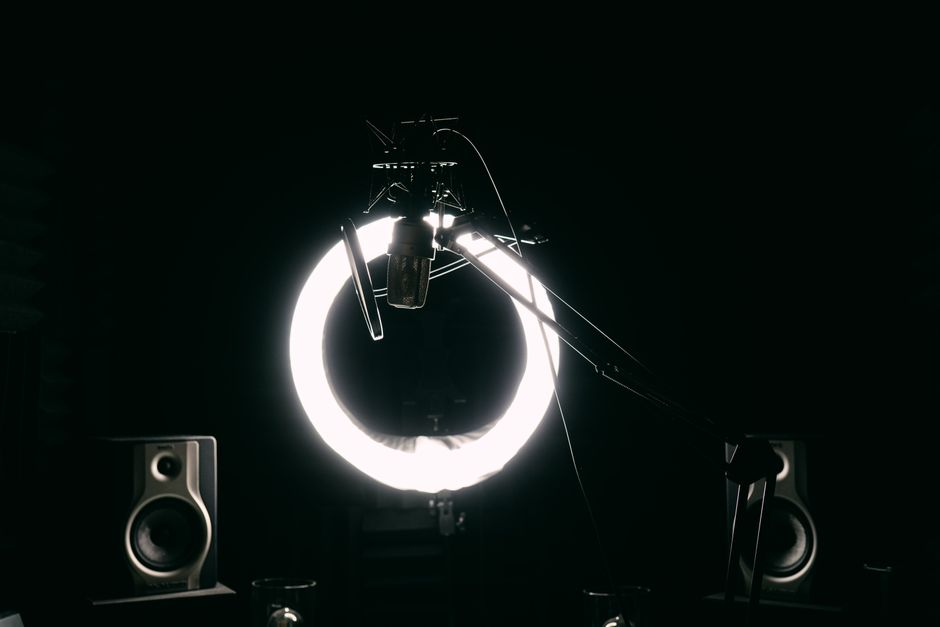 Dark studio room with condenser microphone, ring light, and speakers for recording purposes.